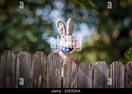 Hasenfigur, die auf einem Holzzaun montiert ist und eine patriotische Gesichtsmaske vor einem Bokeh-Hintergrund trägt. Stockfoto