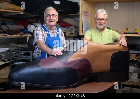 19. August 2021, Baden-Württemberg, Münsingen: Schuhmeister Matthäus Jörg (l.) und Johannes Schunk stehen in Jörg's Werkstatt hinter einem überdimensionalen Schuh. Jörg und Schunk haben den Schuh in der Größe 240 mit Hilfe eines Zimmermanns angefertigt. (To dpa 'Made in Münsingen - zwei Schuhmacher produzieren Schuhe der Größe 240') Foto: Marijan Murat/dpa Stockfoto