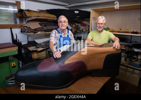 19. August 2021, Baden-Württemberg, Münsingen: Schuhmeister Matthäus Jörg (l.) und Johannes Schunk stehen in Jörg's Werkstatt hinter einem überdimensionalen Schuh. Jörg und Schunk haben den Schuh in der Größe 240 mit Hilfe eines Zimmermanns angefertigt. (To dpa 'Made in Münsingen - zwei Schuhmacher produzieren Schuhe der Größe 240') Foto: Marijan Murat/dpa Stockfoto