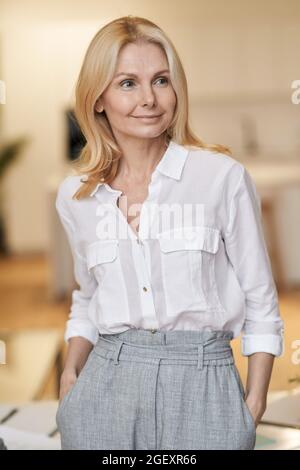 Elegante, reife Geschäftsfrau mit blondem Haar, das beim Stehen im Haus zur Seite schaut Stockfoto