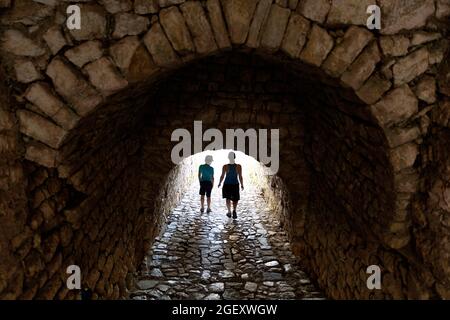 Mutter und Sohn, die durch einen Steinbogengang gehen, Blick auf die Straße im Dorf Vuno, mit einfallenden Sonnenstrahlen, sahen den Bogen, Südalbanien Stockfoto