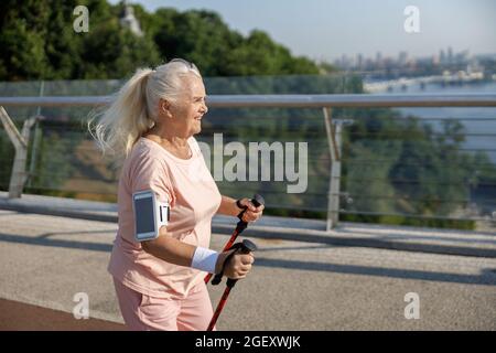 Ältere Frau mit Smartphone praktiziert Nordic Walking auf leerem Steg Stockfoto