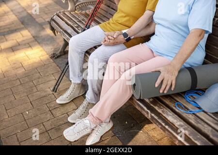 Reifes Paar mit Sportausrüstung schließt sich den Händen an, die in der Nähe des Parks auf der Bank sitzen Stockfoto