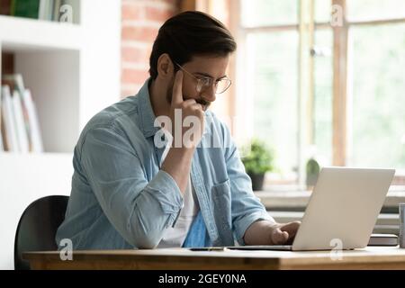 Ein ernsthafter Mann sitzt am Schreibtisch und schaut auf den Laptop, um E-Mails zu lesen Stockfoto