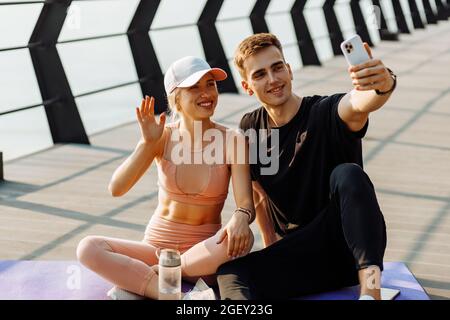 Attraktive junge Sportler, Männer und Frauen, die sich nach dem Training beim Selfie auf dem Mobiltelefon ausruhen, sitzen auf der Fitnessmatte im Freien am Pier Stockfoto