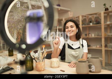 Positive junge Künstlerin demonstriert Werkzeuge für handgemachte Keramik während der Dreharbeiten Video für soziale Netzwerke in professionellen Workshop Stockfoto