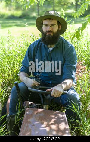 Ein hübscher kaukasischer Rancher, der auf einem alten Grasschneider auf dem Feld sitzt Stockfoto