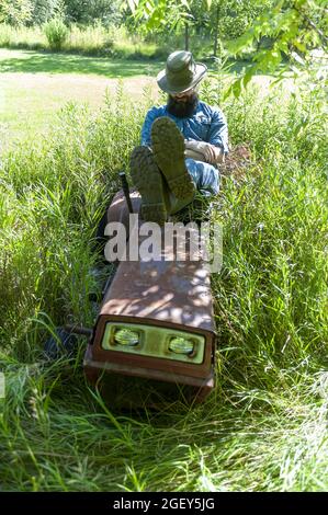 Ein hübscher kaukasischer Rancher, der auf einem alten Grasschneider auf dem Feld sitzt Stockfoto