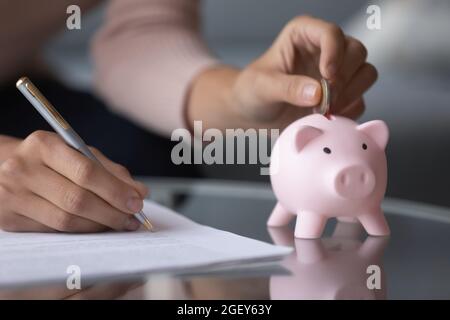 Weibliche Hand legt Münzen in Sparschwein schreibt Einkommensausgaben Stockfoto