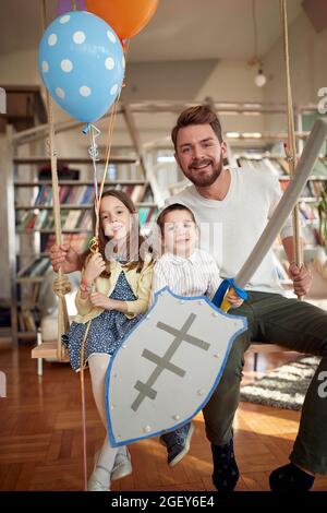 Ein glücklicher Vater sitzt auf der Schaukel in familiärer Atmosphäre zu Hause mit seinen Kindern und posiert für ein Foto. Familie, Freizeit, zusammen Stockfoto