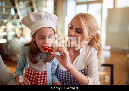 Eine kleine Tochter, die das Gemüse ihrer Mutter verkostet, während sie gemeinsam zu Hause in fröhlicher Atmosphäre ein Essen zubereitet. Familie, Freizeit, gemeinsam Stockfoto