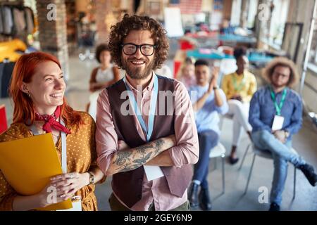 Junge Rothaarige und bärtige Männer mit lockigen Haaren stehen zusammen und posieren für ein Foto vor einer Gruppe von Kollegen im Hintergrund, lächelnd. Stockfoto