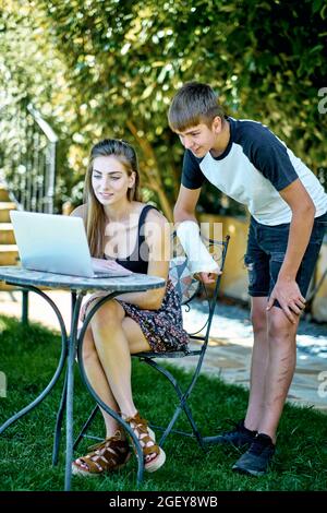 Porträt einer jungen kaukasischen Frau mit ihrem jüngeren Bruder, die draußen in einem Garten posiert, mit einem Laptop, der im Internet nach Informationen sucht. Lifestyle-Conce Stockfoto