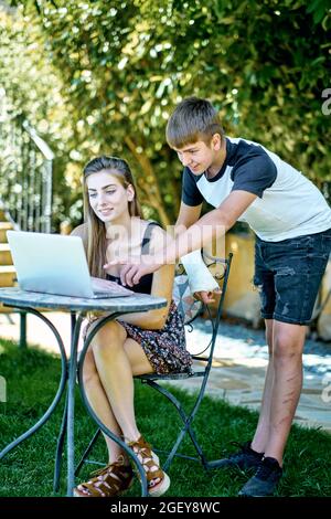 Porträt einer jungen kaukasischen Frau mit ihrem jüngeren Bruder, die draußen in einem Garten posiert, mit einem Laptop, der im Internet nach Informationen sucht. Lifestyle-Conce Stockfoto