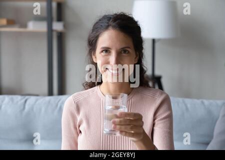 Die junge lateinische Frau sitzt auf dem Sofa und hält ein Glas Wasser Stockfoto
