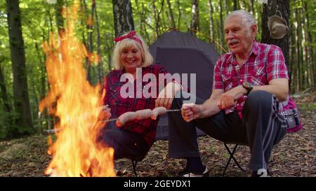 Alte glückliche Großeltern, die sich beim Camping im Wald ausruhen und Würstchen über dem Lagerfeuer braten Stockfoto