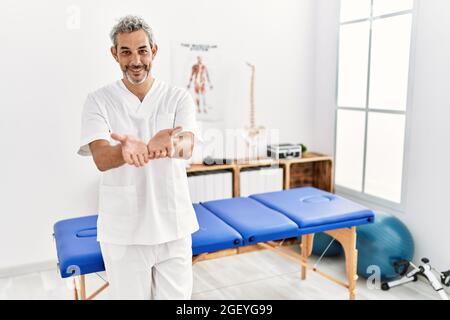 hispanischer Therapeut im mittleren Alter, der in der Schmerzheilungsklinik arbeitet, lächelt mit Handflächen, die zusammen Geste empfangen oder geben. Halt und Schutz Stockfoto