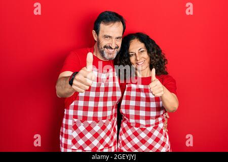 Mittelalterpaar hispanische Frau und Mann tragen Kochschürze Genehmigung tun positive Geste mit der Hand, Daumen hoch lächelnd und glücklich für den Erfolg. wi Stockfoto