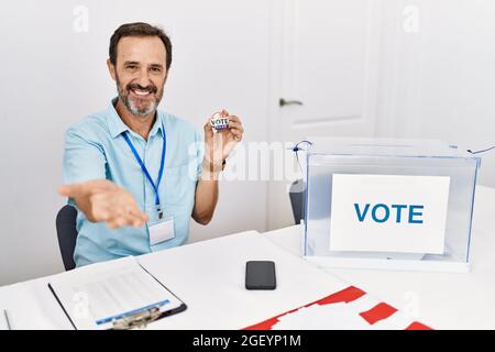 Mann mittleren Alters mit Bart sitzend bei Stimmzettel Ich Stimmzettel Abzeichen lächelnd freundlich bietet Handfläche, die Unterstützung und Akzeptanz. Stockfoto