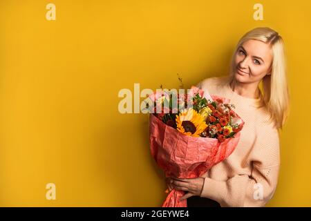 Die junge Frau lächelt und hält einen großen und schönen Herbststrauß mit Sonnenblumen, Chrysanthemen und Rosen auf gelbem Hintergrund mit Kopierraum Stockfoto