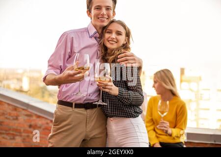 Glückliches junges Paar in Liebe umarmt sich gegenseitig auf der Terrasse des Hauses, hält Gläser Weißwein, lächelt und posiert vor der Kamera, Mann und ihre geliebte Frau Stockfoto