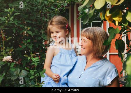 Glückliche liebevolle Oma mittleren Alters hält kleine süße Vorschulengroßtochter, während sie im Garten am Sommertag steht, Großmutter mit kleinem Mädchen verbringen Stockfoto