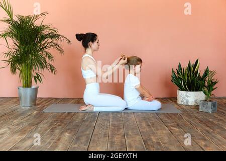 Junge blonde Mädchen bekommen ihre langen Haare geflochten von der Mutter vor dem Sporttraining, während sie zu Hause oder im Fitnessstudio auf dem Boden sitzen Stockfoto