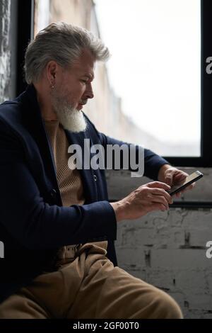 Fokussierter Mann mittleren Alters in Business Casual Wear mit seinem Smartphone, während er an einem Fenster im modernen Loft-Interieur sitzt Stockfoto