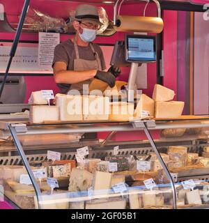 Limoux Aude France 08.20.21 der Mann mit Gesichtsmaske und Handschuhen wickelt eine Scheibe Käse in braunes Papier. Die gekühlte Glastheke zeigt eine große Auswahl Stockfoto