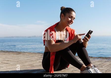 Außenaufnahme einer attraktiven Sportlerin, die in der Nähe des Meeres eine Pause macht, auf einem hölzernen Pier sitzt und ein Mobiltelefon benutzt Stockfoto