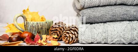 Herbstlich gelbe Ahornblätter, Tannenzapfen, Kastanien und eine Tasse Tee neben einem Stapel von gestrickten gemütlichen warmen Winterpullover in mehreren Grautönen auf einem Stockfoto