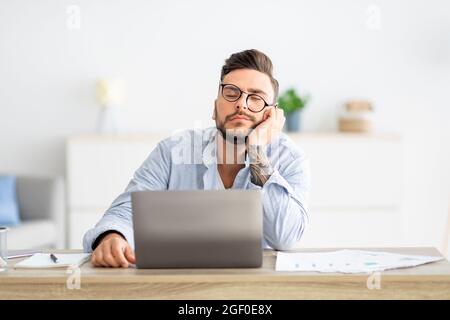 Überarbeiteter junger freiberuflicher Mann, der am Arbeitsplatz sitzt und schläft, beim Arbeiten am Laptop, beim Kopieren des Speicherplatzes schlaft Stockfoto