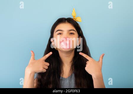 Lustige indische Teenager-Mädchen Blasen Blase aus Zucker frei Kaugummi, zeigt auf sie über blauen Studio-Hintergrund Stockfoto