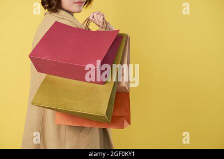 Beschnittenes Bild einer jungen Frau, die viele Einkaufstaschen hält und sich zur Kamera zurückwendet, isoliert auf Gelb Stockfoto
