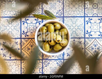 Kleine weiße Schale mit einem Snack aus grünen Oliven auf einem Tisch mit traditionellen mediterranen blauen Kacheln unter einem Olivenbaum Stockfoto