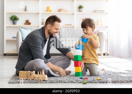 Spielzeit. Fröhlicher, liebevoller Vater, der mit seinem kleinen Sohn bunte Blöcke spielt, Junge, der im Inneren des Hauses einen großen Turm baut Stockfoto