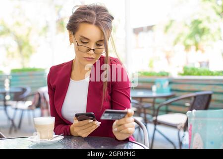 Eine nachdenkliche Frau mit Smartphone und Kreditkarte sitzt in einem Sommercafe. Online-Shopping-Konzept. Stockfoto