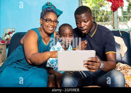 Die schöne afrikanische Familie, die im Sessel sitzt, schaut sich zusammen ein Video auf dem digitalen Tablet an, während sie alle glücklich sind Stockfoto