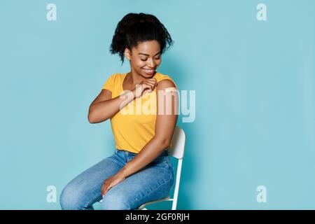 Lächelnde schwarze Frau, die gegen Covid geimpft wird, zeigt Arm mit Bandhilfe auf blauem Hintergrund, freier Platz Stockfoto