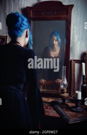 Drinnen Porträt von schönen Goth Mädchen in schwarzem Kleid. Blauhaarige Gothic Lady, die in den alten schmutzigen Spiegel schaut. Vintage-Style Stockfoto