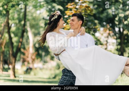Glückliches Hochzeitspaar, der Bräutigam bewundert die glückliche Braut, hält die Braut in den Armen und wirbelt sie, an einem sonnigen Sommertag im Park, fröhlich zur Hochzeit Stockfoto