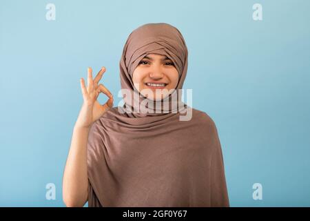 Fröhliche muslimische Teenager-Mädchen in Hijab zeigt okay Geste auf blauen Studio-Hintergrund Stockfoto