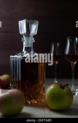 Vintage-Glas mit hausgemachten Craft Calvados mit drei Äpfeln und zwei Gläsern auf Marmortisch in dunkler Stimmung. Stockfoto