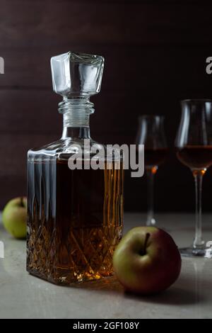 Vintage-Glas mit hausgemachten Calvados mit zwei Äpfeln und zwei Gläsern auf Marmortisch in dunkler Stimmung. Stockfoto