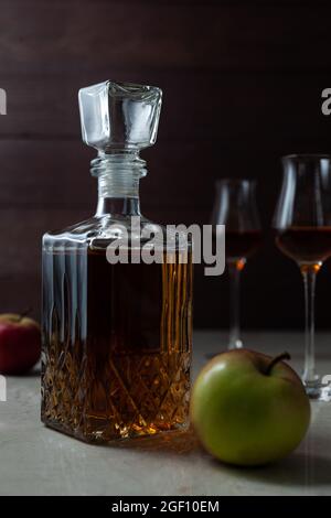 Vintage-Glas mit hausgemachten Craft Calvados mit zwei Äpfeln und zwei Gläsern auf Marmortisch in dunkler Stimmung. Stockfoto