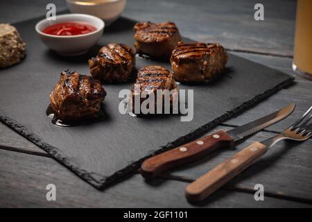 Gegrillte Schweinemedaillons mit Ketchup auf Steinbrett auf dunklem Holztisch. Stockfoto