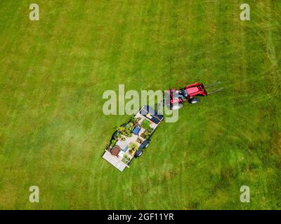 Ein roter Traktor zieht an einem sonnigen, klaren Sommertag einen Trailor mit Pflanzen auf einem grünen Grasfeld. Luftperspektive mit Blick nach unten. Stockfoto