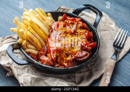 Currywurst mit pommes in schwarzer Pfanne auf Bastelpapier auf Holztisch. Stockfoto