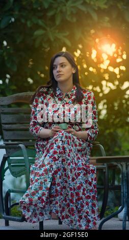 Schöne lateinische Frau in einem langen Kleid mit floralem Design sitzt im Garten mit einer Tasse Kaffee in den Händen während des Nachmittags mit der Sonne Stockfoto