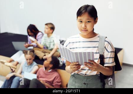 Waist-up-Porträt eines jungen Schulmädchen, das Rucksack trägt und die Kamera anschaut, während es in der Schulhalle steht und Platz kopiert
